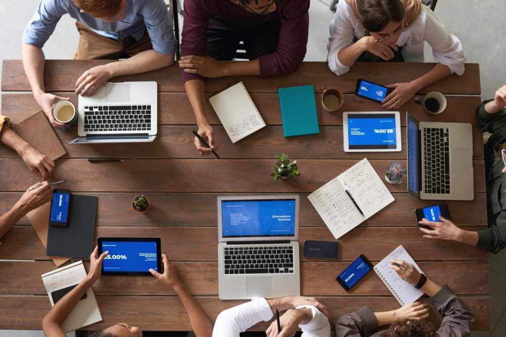 7 Best Mobile App Makers in 2022 To Make Your Own Mobile App - an overhead view of a wooden plank table with a load of laptops on it with people using them and taking notes on paper pads